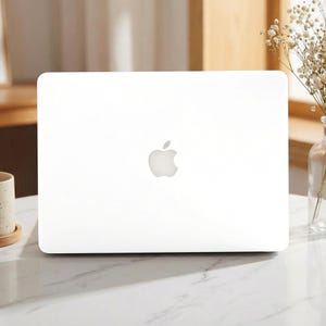 May include: A closed laptop with a solid white exterior shell and a centred silver Apple logo. The device features a smooth, matte finish on the lid, presenting a clean and uniform appearance across the entire surface of the computer casing.