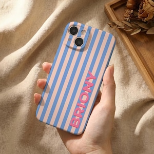 Puede incluir: Funda de tel&eacute;fono a rayas verticales azules y rosas, con la palabra "BRIONY" en rosa. La funda tiene un recorte para doble c&aacute;mara. La funda se sostiene en una mano.