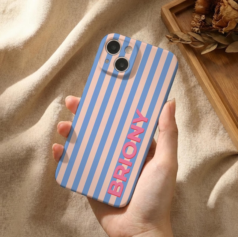 Puede incluir: Funda de tel&eacute;fono a rayas verticales azules y rosas, con la palabra "BRIONY" en rosa. La funda tiene un recorte para doble c&aacute;mara. La funda se sostiene en una mano.
