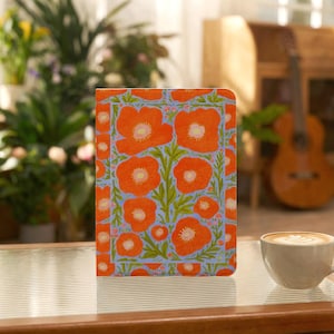 Puede incluir: Una funda rectangular para tableta con un patrón vibrante de flores de amapola naranjas con centros rosa claro y tallos verdes sobre un fondo azul claro, exhibida en posición vertical sobre una superficie junto a una taza de café de cerámica blanca.