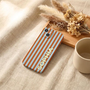 Puede incluir: Funda de tel&eacute;fono con un patr&oacute;n de rayas naranjas y celeste, con el nombre "SOPHIA" en letras doradas. La funda tiene dos lentes de c&aacute;mara. Flores secas y una taza de cer&aacute;mica en el fondo, sobre una superficie beige.