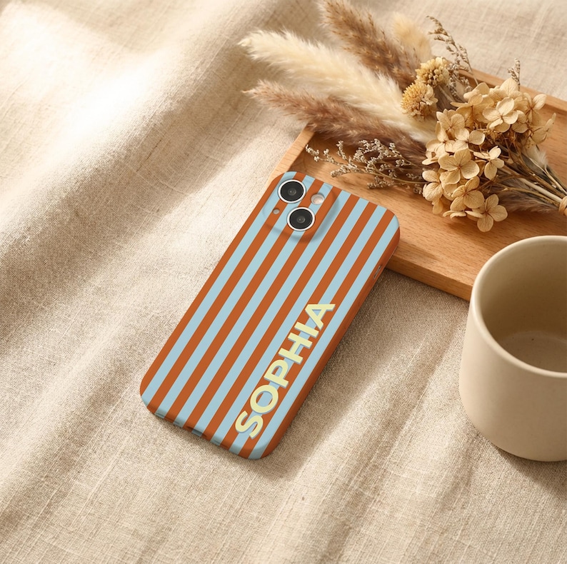 Puede incluir: Funda de tel&eacute;fono con un patr&oacute;n de rayas naranjas y celeste, con el nombre "SOPHIA" en letras doradas. La funda tiene dos lentes de c&aacute;mara. Flores secas y una taza de cer&aacute;mica en el fondo, sobre una superficie beige.