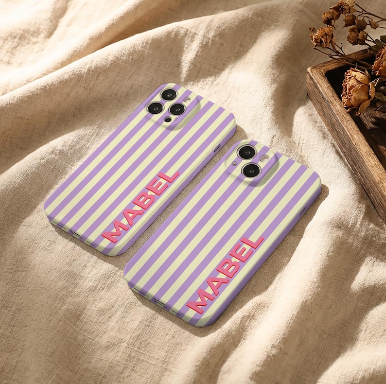 Puede incluir: Dos fundas de tel&eacute;fono con un patr&oacute;n de rayas verticales en lavanda y crema. La palabra "MABEL" est&aacute; impresa en rosa en cada funda. Las fundas est&aacute;n sobre una tela beige.