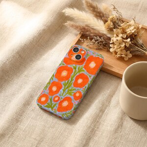Puede incluir: Una funda rectangular para smartphone con un patr&oacute;n vibrante de flores de amapola de color naranja brillante con centros amarillos y tallos verdes sobre un fondo azul b&iacute;garo claro. La funda tiene dos aberturas circulares para las lentes.