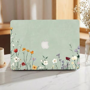 Könnte beinhalten: Eine hellgrüne Laptop-Hülle mit einem Blumendesign aus bunten Wildblumen. Das Gehäuse hat ein weißes Apple-Logo in der Mitte. Die Blumen sind rot, orange, weiß und lila.