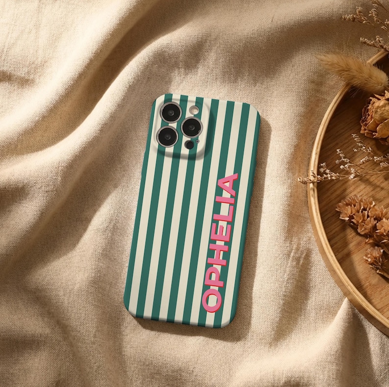 Puede incluir: Una funda de tel&eacute;fono con un patr&oacute;n de rayas verticales en verde azulado y blanco roto. La palabra "OPHELIA" est&aacute; impresa verticalmente en rosa. La funda tiene un recorte para la c&aacute;mara y est&aacute; sobre una tela beige.
