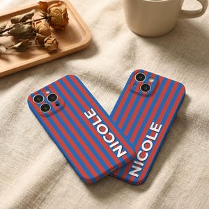 Puede incluir: Dos fundas de tel&eacute;fono con un dise&ntilde;o de rayas azules y rojas. La palabra "NICOLE" est&aacute; impresa verticalmente en blanco en cada funda. Las fundas est&aacute;n sobre una superficie beige.