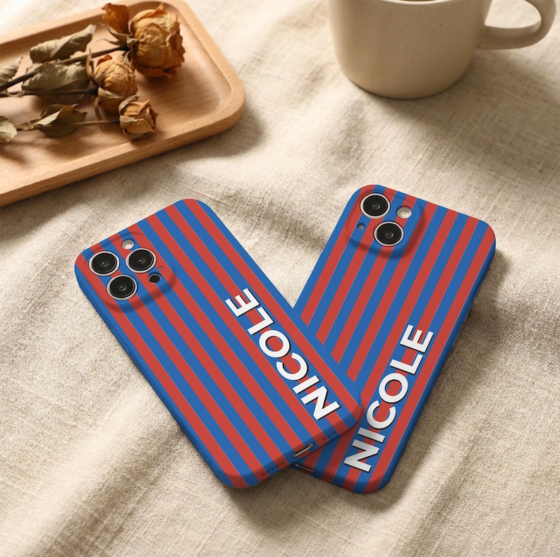 Puede incluir: Dos fundas de tel&eacute;fono con un dise&ntilde;o de rayas azules y rojas. La palabra "NICOLE" est&aacute; impresa verticalmente en blanco en cada funda. Las fundas est&aacute;n sobre una superficie beige.
