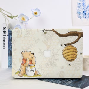 May include: A beige laptop case with a cartoon illustration of Winnie the Pooh and a honey pot labelled "Hunny", alongside a beehive. The case has a honeycomb pattern and a tree branch with bees.