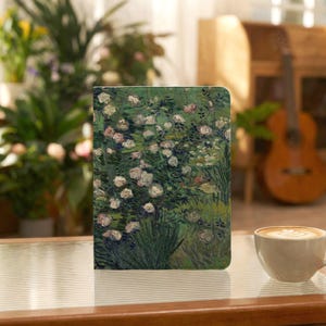 Puede incluir: Una funda rectangular para tableta con una reproducción impresa de una pintura de rosas blancas sobre un fondo de follaje verde oscuro. La funda está colocada en posición vertical sobre una superficie junto a una taza de cerámica blanca con café.