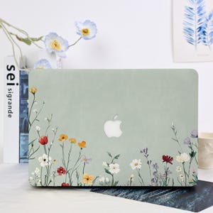 Könnte beinhalten: Eine hellgrüne Laptop-Hülle mit floralem Design. Die Hülle zeigt ein weißes Apple-Logo und einen Rand aus bunten Wildblumen in Rot, Gelb, Orange, Weiß und Lila. Die Blumen sind vor einem hellgrünen Hintergrund platziert.