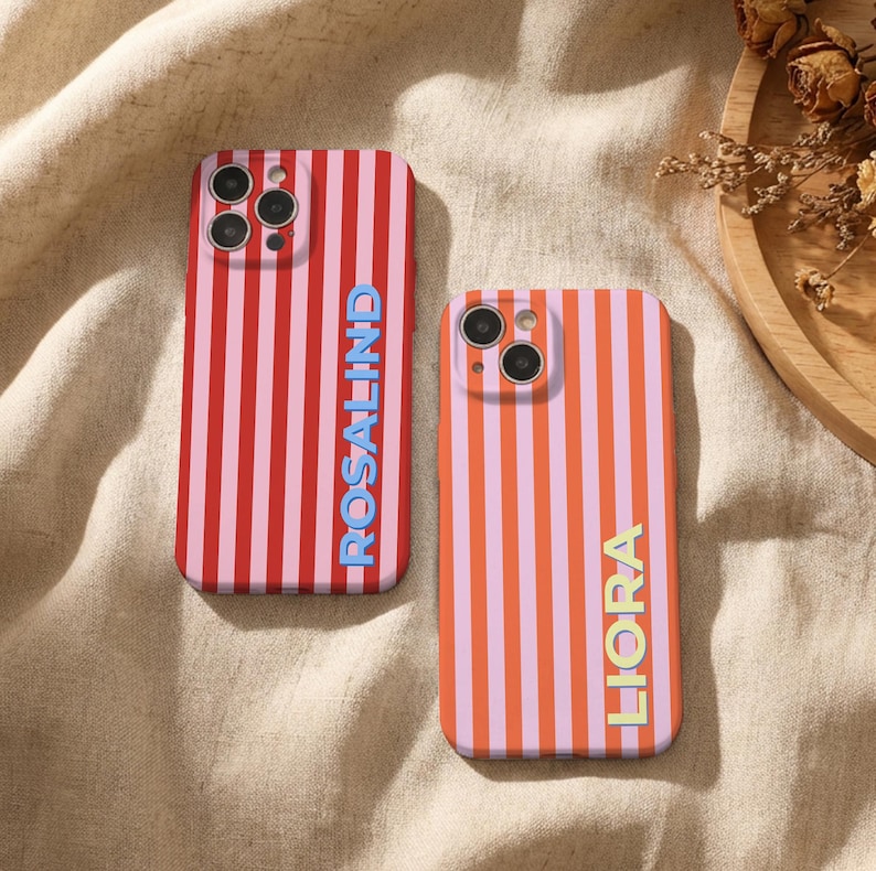 Puede incluir: Dos fundas de tel&eacute;fono con rayas verticales. Una funda es roja y rosa con el nombre "ROSALIND" en azul. La otra funda es naranja y rosa con el nombre "LIORA" en amarillo. Ambas fundas tienen un recorte para la c&aacute;mara.