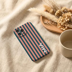 Puede incluir: Funda de tel&eacute;fono con rayas verticales en verde azulado y coral. El nombre "MIA" est&aacute; en letras blancas. La funda est&aacute; sobre una tela beige, con una bandeja de madera con flores secas y una taza beige en el fondo.