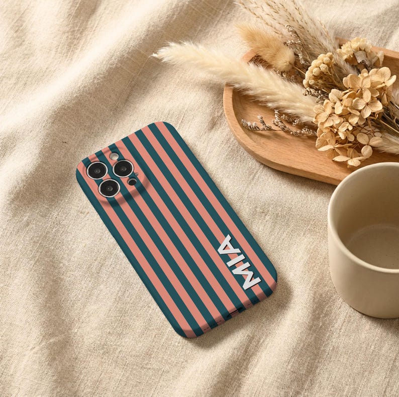 Puede incluir: Funda de tel&eacute;fono con rayas verticales en verde azulado y coral. El nombre "MIA" est&aacute; en letras blancas. La funda est&aacute; sobre una tela beige, con una bandeja de madera con flores secas y una taza beige en el fondo.
