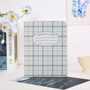 Puede incluir: Un cuaderno de composición con un patrón de cuadros azules y blancos. La portada presenta una etiqueta ovalada con las palabras "composition book" y líneas para escribir. El cuaderno se muestra con un libro y flores en el fondo.