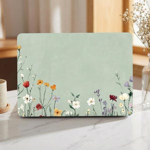 Könnte beinhalten: Eine rechteckige Laptop-Hülle mit hellgrünem Hintergrund und floralem Design. Das Design zeigt bunte Wildblumen in Rot-, Orange-, Weiß- und Lilatönen am unteren Rand. Die Hülle ist für einen Laptop konzipiert.