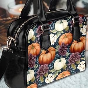 May include: A black leather handbag with a shoulder strap and a floral print featuring pumpkins, white flowers, and berries. The print is on a dark blue background.