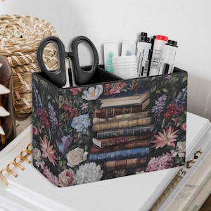 Op de afbeelding: Een zwarte bureau-organizer met een bloemen- en boekontwerp. De organizer heeft twee compartimenten en is gevuld met kantoorbenodigdheden, waaronder een schaar, pennen en stiften.
