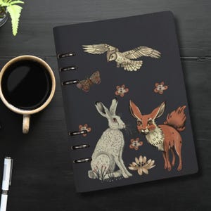 Könnte beinhalten: Ein schwarzes Spiralnotizbuch mit einem dunkelblauen Einband, auf dem ein weißes Kaninchen, ein orangefarbener Fuchs, ein Schmetterling und eine Eule abgebildet sind. Die Tiere sind von kleinen weißen Blumen umgeben.
