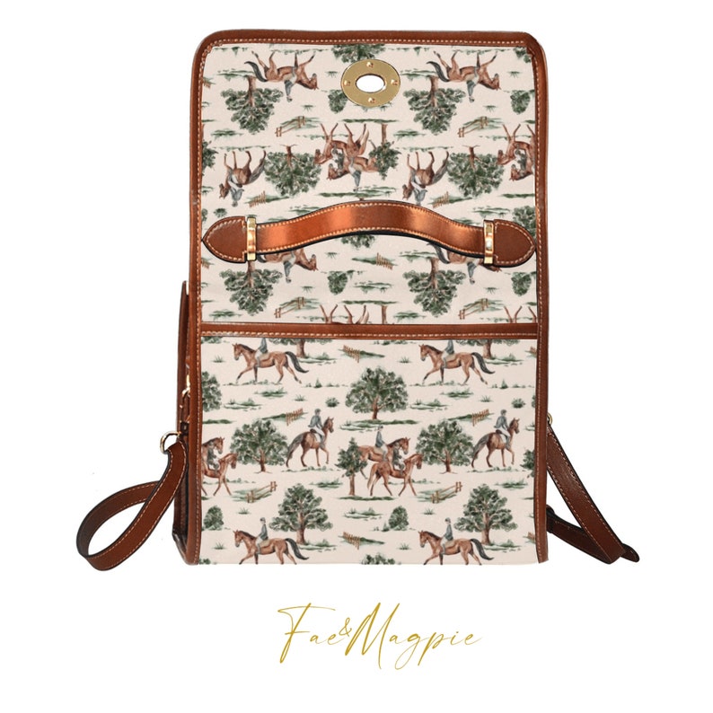 K&ouml;nnte beinhalten: Eine braune Ledertasche mit einem wei&szlig;en Canvas-Druck von Pferden und Reitern, die &uuml;ber Hindernisse springen. Die Tasche hat einen oberen Griff und einen langen Schultergurt. Die Tasche wird mit einer Messing-Schnalle geschlossen. Die Tasche hat den Text "Fae & Magpie" auf der Unterseite gedruckt.