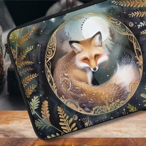 May include: A laptop sleeve with a dark blue background featuring a stylised illustration of a fox with a bushy tail. The fox is surrounded by a gold-coloured circular frame with intricate details. The background is decorated with gold-coloured leaves and a moon.