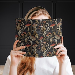 Può includere: Un taccuino nero con un motivo colorato di funghi e felci. Il taccuino è tenuto da una persona.