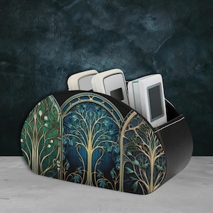 Könnte beinhalten: Ein schwarzer Leder-Fernbedienungsständer mit einem dekorativen Design mit drei Bäumen mit goldenen Ästen und blauen und grünen Blättern. Der Ständer hat drei Fächer für Fernbedienungen.