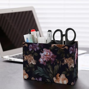 May include: A black desk organiser with a floral pattern featuring pink, white, and purple flowers. The organiser has four compartments and is filled with pens, markers, and scissors.