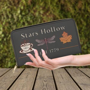 May include: A dark gray rectangular zip-around wallet featuring the text Stars Hollow, a graphic of a coffee cup labeled Luke's, a dragonfly, an orange maple leaf, and the text EST 1779. The wallet has a gold-toned zipper closure.