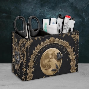 Puede incluir: Un organizador de escritorio negro y dorado con un diseño de luna y flores. El organizador tiene tres compartimentos y es perfecto para guardar bolígrafos, lápices, tijeras y otros artículos de oficina.