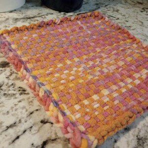 May include: A woven potholder in shades of orange, pink, and yellow. The square potholder has a braided edge and a small loop for hanging. The weave pattern is visible, and the colors blend together.