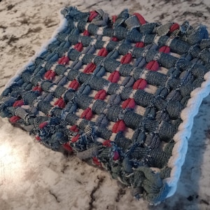 May include: A square, handmade pot holder with a woven design. The pot holder is made of blue denim fabric with pink accents and a white border. The woven pattern creates a textured surface.