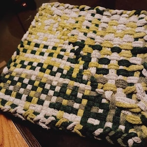 May include: A square, woven hot pad in shades of green, yellow, and off-white. The handmade pot holder features a loop for hanging and a textured, checkered pattern. The woven design provides heat resistance for kitchen use.