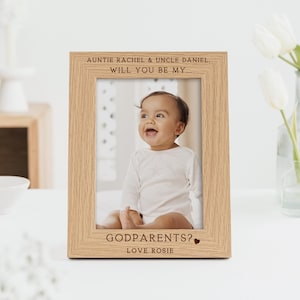 May include: A light oak picture frame with a photograph of a smiling baby. The frame has engraved text at the top and bottom, including "AUNTIE RACHEL & UNCLE DANIEL. WILL YOU BE MY... GODPARENTS? LOVE ROSIE."