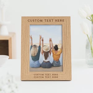 Peut inclure: Un cadre photo en ch&ecirc;ne clair avec une photo de trois personnes les bras lev&eacute;s. Le cadre porte le texte "CUSTOM TEXT HERE" en haut et en bas. Un vase de tulipes blanches est en arri&egrave;re-plan.
