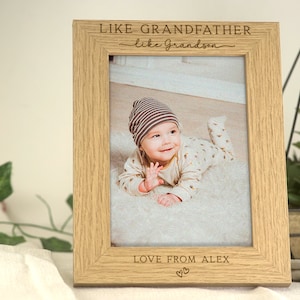 May include: A light wood photo frame with a baby in a striped hat and a white onesie. The frame has the text "Like Grandfather, Like Grandson" and "Love from Alex" with a heart symbol.