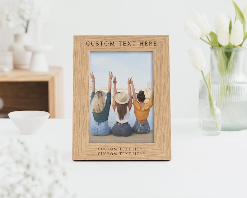 Peut inclure: Un cadre photo en ch&ecirc;ne clair avec une photo de trois personnes les bras lev&eacute;s. Le cadre porte le texte "CUSTOM TEXT HERE" en haut et en bas. Un vase de tulipes blanches est en arri&egrave;re-plan.