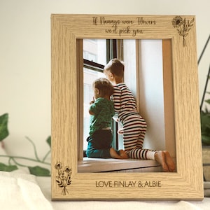 May include: A wooden photo frame with a light brown finish. The frame has a white mount and a photo of two children looking out a window. The frame is engraved with the text "If Nannys were Flowers we'd pick you" and "LOVE FINLAY & ALBIE".