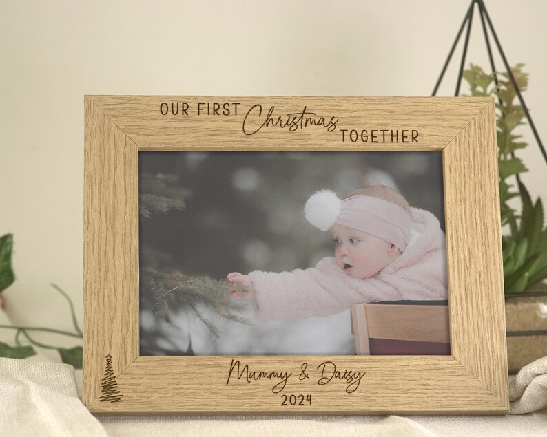 Pu&ograve; includere: Una cornice per foto in legno con il testo "Our First Christmas Together" e "Mommy & Daddy 2024". La cornice contiene una foto di una bambina vestita di rosa.