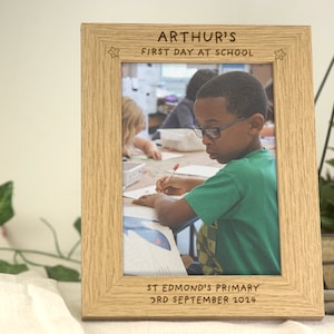 May include: A wooden photo frame with a personalised inscription for a child's first day of school. The inscription reads "Arthur's First Day at School" with stars on either side. The inscription at the bottom reads "St Edmond's Primary 3rd September 2024". The frame contains a photo of a young boy in a green shirt sitting at a desk in a classroom.