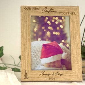Pu&ograve; includere: Una cornice per foto in legno con il testo "Our First Christmas Together" e "Mommy & Daddy 2024" inciso su di essa. La cornice contiene una foto di un bambino che indossa un cappello di Babbo Natale.