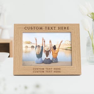 Peut inclure: Un cadre photo en bois brun clair avec le texte "CUSTOM TEXT HERE" grav&eacute; en haut et en bas. Le cadre contient une photo de trois personnes les bras lev&eacute;s. Un vase de fleurs blanches est en arri&egrave;re-plan.