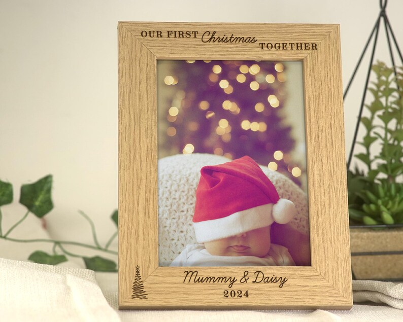 Pu&ograve; includere: Una cornice per foto in legno con finitura marrone chiaro. La cornice presenta un'incisione di un albero di Natale e il testo "Our First Christmas Together" in alto. La cornice ha anche il testo "Mummy & Daisy 2024" in basso. La cornice contiene una foto di un bambino che indossa un cappello rosso di Babbo Natale.