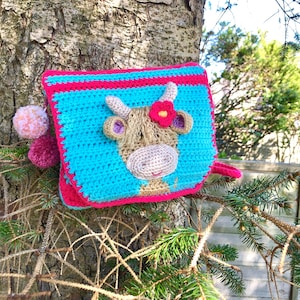 May include: A crocheted cow-shaped pouch with a pink flower on its head. The pouch is blue with pink and red stripes. It is hanging from a tree branch.