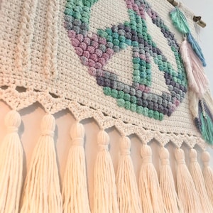 May include: A crocheted wall hanging with a peace sign design in shades of pink, green, and purple. The peace sign is surrounded by a white crocheted border and the hanging has a fringe of white tassels.