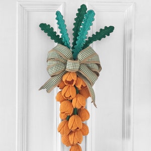 Op de afbeelding: Een handgemaakte gehaakte deurhanger in de vorm van een wortel. De wortel heeft oranje tulpvormige bloemen, groene stelen en een decoratieve strik met een ruitpatroon. De deurhanger is ontworpen om aan een deur te worden gehangen.