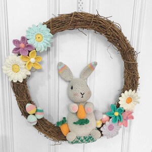 Puede incluir: Un conejito de crochet sosteniendo una zanahoria se sienta en una corona de ramas decorada con flores y huevos de Pascua de crochet.