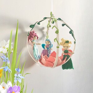 May include: A white wooden hoop mobile with three crocheted fairies and a pink crocheted flower. The fairies are wearing pink, blue, and green dresses. The mobile is hanging from a white ceiling.