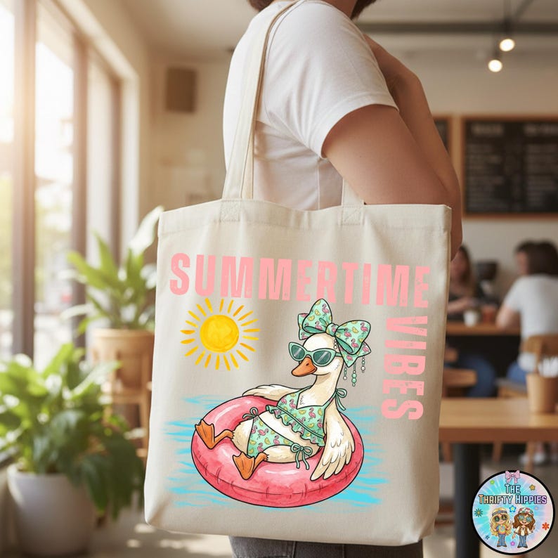 Puede incluir: Bolso tote color crema con las palabras "SUMMERTIME VIBES" en rosa. El bolso presenta un ganso de dibujos animados con gafas de sol y un traje de ba&ntilde;o, flotando en un anillo inflable rosa. Tambi&eacute;n se incluye un gr&aacute;fico de sol amarillo.