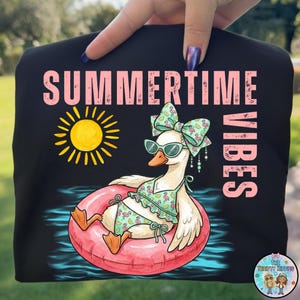 Puede incluir: Camiseta negra con la frase "SUMMERTIME VIBES" en rosa. Un dibujo de un ganso con gafas de sol y bikini flota en un flotador rosa. Tambi&eacute;n hay un sol amarillo.
